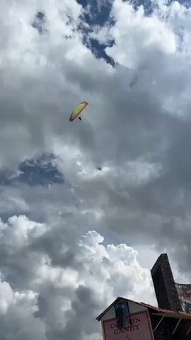 The video shows a person paragliding against a cloudy sky. The paraglider is colorful, with yellow, red, and green sections. A person is suspended beneath the paraglider. In the foreground, the top of a building with a sign that says "COTTON CANDY" is visible. To the right of the building, a tall, dark, stone structure stands. The sky is filled with large, dramatic clouds, with patches of blue sky visible. The overall impression is one of adventure and outdoor activity.