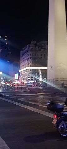 At night, a street scene reveals the Obelisco de Buenos Aires monument in Argentina. A motorcycle with a rear storage container stands at a crosswalk, its brake light illuminated. After a brief pause, the motorcycle proceeds. A group of people, dressed in casual attire, cross the street in the zebra crossing. The setting is urban, with illuminated buildings and large electronic billboards displaying light and advertisements. The street is wet, reflecting the city lights, and a few cars are visible in the background.
