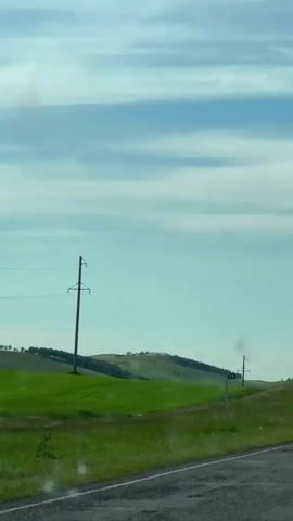The video shows a view from a vehicle moving along a paved road. The road runs along the right side of the frame, with a grassy shoulder marked by white posts. To the left of the road is a vast, undulating green field, likely farmland. In the distance, rolling hills are visible, dotted with what appear to be small buildings or structures. Power lines stretch across the landscape, supported by utility poles. The sky above is a clear blue with wispy white clouds. The overall impression is of a rural, open landscape under a daytime sky.