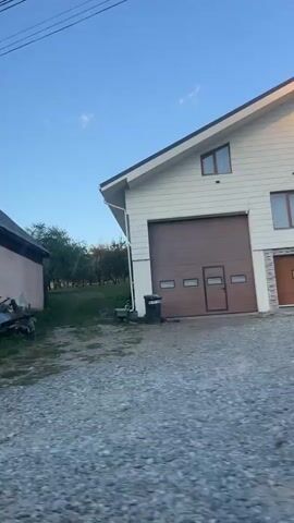 The video shows a moving vehicle's perspective of passing by several houses. The first house is pink with a tiled roof and a white and grey fence. The second house is beige with an arched entryway and a white balustrade. The third scene shows a wooden fence with a black car parked behind it, next to a small wooden building. The final scene is of a beige house with a dark roof and intricate white detailing around the windows. The sky is blue with some clouds, and there are power lines visible throughout the video.