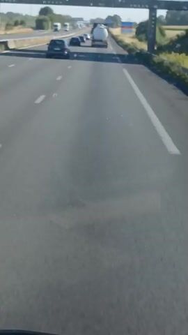 A dash cam view from a vehicle on a highway shows traffic ahead. The road is paved and has lane markings. Several cars and trucks are visible on the highway, including a white tanker truck. The surrounding environment appears to be rural with trees and vegetation. The sky is clear and the sun is shining.