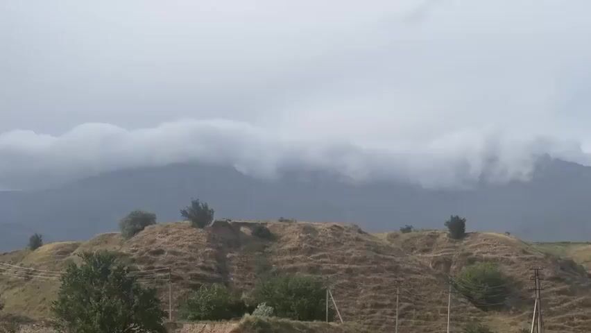 The video shows a landscape with rolling hills in the foreground and a mountain range in the background. The hills are covered in dry, yellow grass and sparse green bushes. Several trees are scattered across the hills. In the background, the mountain range is partially obscured by a thick, low-lying cloud formation that stretches across the peaks. The sky above is overcast with grey clouds. There are power lines with utility poles visible on the right side of the frame. The scene appears to be a rural or natural environment, possibly a mountainous region under cloudy weather.