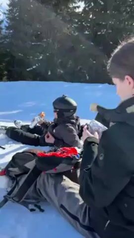 Two people are sitting in the snow, wearing winter gear, and appear to be taking a break. One person is wearing a black jacket and a helmet with goggles, holding a bottle and a wrapper. The other person is wearing a dark jacket and also has a wrapper. A gray backpack is next to them. The camera pans up to reveal a scenic view of a snowy mountain range under a clear blue sky, with trees dotting the landscape. There are ski slopes visible with markers.