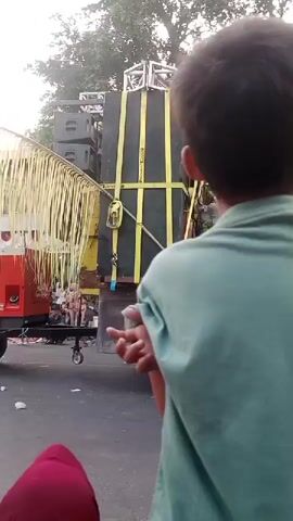 The video opens with a close-up of the back of a young person wearing a light blue t-shirt. The camera then pans right to reveal a large sound system mounted on the back of a truck. The sound system is tall and black, with yellow straps holding it in place. There are also some decorative palm fronds hanging from the truck. A person is riding a black motorcycle in front of the truck, and another person is sitting behind them. The camera then focuses on a young child standing to the right of the frame, who appears to be covering their mouth with their hands. In the background, there are trees and a building. The overall scene suggests an outdoor event or gathering.