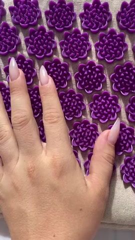 A hand with long, tapered fingernails, painted with a sheer, milky polish, rests on a mat covered in rows of small, purple, flower-shaped acupressure points. The hand is positioned in the center of the frame, with the fingers extended and gently pressing down on the mat. The mat itself is a neutral beige fabric, and the purple acupressure points are evenly spaced in a grid pattern. The lighting is bright, casting soft shadows and highlighting the texture of the mat and the smooth surface of the acupressure points. The video is a close-up shot, focusing solely on the hand and the mat.
