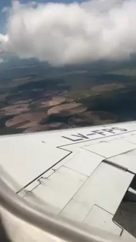 From the window of an airplane, a view of the land below is seen. The plane wing is white with black lettering reading “LV-FPS” and divides the frame horizontally. Below the wing, a landscape of rural farmland is visible. Patches of green and brown land are bisected with circular irrigation patterns. Above the wing, puffy, white clouds are present in the blue sky. The camera is relatively stable.