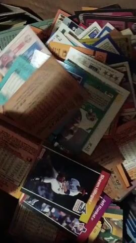 A hand rummages through a box filled with baseball cards. The cards are of various colors and designs. The hand picks up and shuffles through some cards, revealing their fronts and backs. The background is dark and out of focus. The box appears to be cardboard.