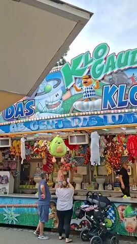 The video shows a carnival game booth. The booth's signage reads "KLO-GAUDI KLO LUTZI". Below the sign, there are multiple toilets lined up, each with a yellow ball placed in front of it. Various stuffed animals and prizes are hanging above and around the toilets. In the foreground, there is a stroller and several people are visible, including a man and a woman looking at the game, and another woman operating the booth. The environment appears to be outdoors, likely at a fair or carnival, with a cloudy sky overhead.