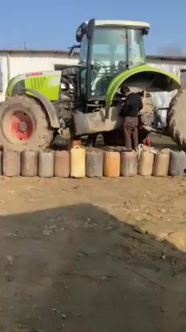 The video shows a man working on a Claas tractor. The tractor, which is lime green and white, has its hood open. The front left wheel of the tractor is jacked up, resting on a line of colorful plastic fuel or liquid containers. The man, wearing a dark shirt and brown pants, is standing near the front of the tractor, appearing to be working on the engine or a related component. The ground is dirt. Behind the tractor is a white building. The sky is blue.