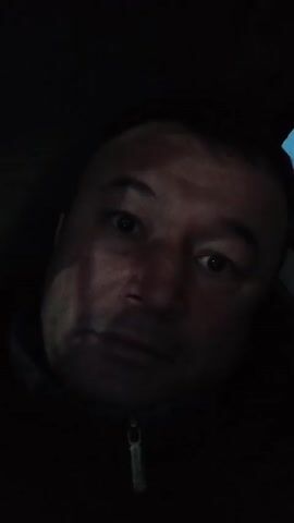 A close-up, low-angle shot of a man's face in dim lighting. He has wide eyes and a slightly concerned expression. He is wearing a dark jacket or coat with a visible zipper. The background is mostly dark, but there is a hint of blue light in the upper right corner, possibly indicating an outside light source.