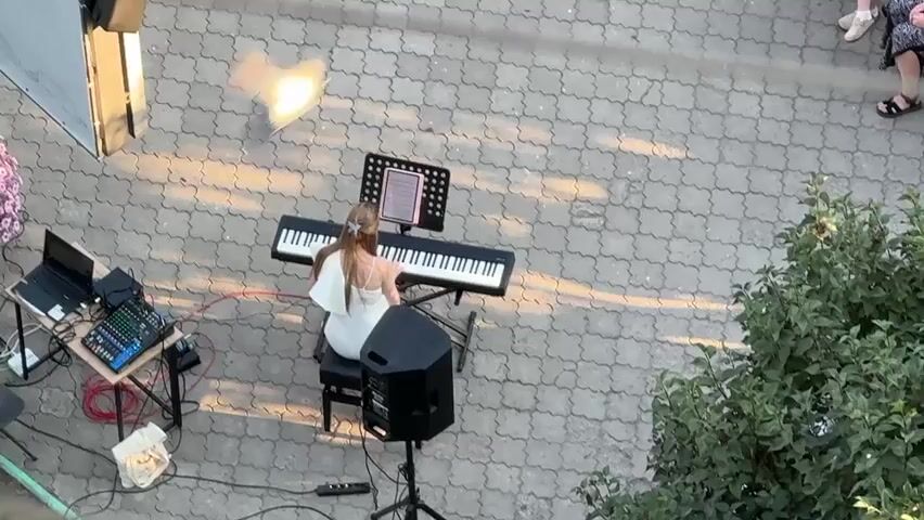 This media file captures an overhead view of a young woman playing a digital piano outdoors. The setting appears to be a paved area, possibly a courtyard or walkway, with a textured pattern of interlocking gray pavers. To the left, a metal stand holds a laptop, a small audio mixer with colorful faders, and various cables, suggesting a performance setup. A black speaker on a stand is positioned in front of the piano.

The woman, dressed in a white sleeveless dress, is seated on a stool and her back is to the camera. Her hair is pulled back. She is focused on the digital piano and the sheet music displayed on a stand above it. Sunlight casts long shadows across the pavement, indicating it is late afternoon or early evening. The presence of lush green foliage to the right suggests a location with vegetation, possibly a garden or park area. In the upper right corner, the legs and lower portion of two individuals are visible, hinting at an audience or passersby.

The overall atmosphere seems calm and focused, with the woman engrossed in her musical performance. The lighting suggests a pleasant, possibly warm, time of day. There is no visible text in the media file.
