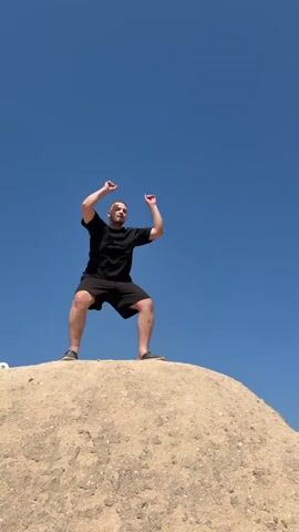 A man is filmed posing outdoors on top of a mound of light brown, rocky earth against a clear, bright blue sky. The man has short light brown hair and a beard, and he is wearing a black t-shirt, black shorts, and dark shoes. He strikes several dynamic poses for the camera: initially, he clenches his fists and bends his knees, then he spreads his arms wide, and finally, he points one arm upwards. The setting suggests a dry, arid environment, consistent with the provided location of Ürgüp, Turkey. The weather is sunny with no clouds. Someone can be heard speaking in the background in Vietnamese, asking, "If I stand here, will I be in the shot, sir?". There is no visible text in the video.