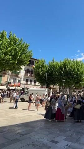 The video shows a sunny, outdoor scene in a town square. The sky is a clear, bright blue with only a few wispy clouds. In the foreground, a group of people dressed in traditional clothing, including long skirts, aprons, and head coverings, are gathered. They appear to be interacting and perhaps preparing for an event or performance. The square is paved with stone and lined with green trees that provide shade. Other people, likely tourists or locals, are milling about, some taking pictures. A white van is parked in the square, and behind it, historic buildings rise up, some adorned with flags. The flags are primarily white with red crosses. The backdrop features mountains visible in the distance. There is a bustling atmosphere, suggesting a festival or celebration is taking place.