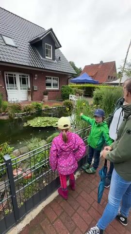 A video captures a boy in a green jacket pointing towards a pond in a residential backyard, while a girl in a pink jacket and yellow hat looks into the pond. The pond features several koi fish swimming and lily pads on the surface. A brick house with a black tiled roof and white window frames stands in the background, with a green canopy visible further back. A metal fence separates the children from the pond.