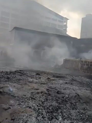 The media file depicts a scene of destruction with smoke billowing from a low-rise building in the foreground. Behind it, a large, multi-story building with visible floors and windows stands, partially obscured by the smoke. The ground in front of the smaller building is covered in debris, ash, and dark soil, suggesting a recent fire or other destructive event. A few scattered objects, including what appears to be a plastic bottle, are visible on the ground. Silhouettes of a few individuals can be seen in the distance, near the smoke, hinting at the presence of people in the area, possibly involved in or observing the event.

The overall atmosphere is somber and chaotic due to the smoke and debris. The lighting suggests it might be daytime, possibly overcast or late afternoon, given the muted light and the sky's appearance. The context provided places this scene in Nairobi, Kenya. The image captures the aftermath of an incident that has caused significant damage, leaving a trail of smoke and devastation in its wake. There is no visible text within the media file.