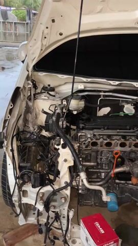 Here's a description of the video:

The video shows the engine compartment of a white car. The engine is exposed, with many parts removed. A red box labeled "GMB" sits on the floor near the engine. The camera pans slowly, showing the intricate layout of the engine and the surrounding components. The overall setting suggests the car is undergoing maintenance or repair.