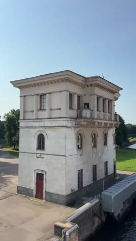 The video opens with a shot of a white building with columns and arched windows, situated next to a body of water. A red door is visible on the left side of the building. The camera then pans to the right, revealing a river or canal with a boat docked alongside the building. The boat has an orange covering on its deck. The camera continues to pan, showing a wider view of the waterway, with lush green trees lining the banks. In the distance, more structures and a dam-like feature can be seen across the water. The sky is clear and blue, indicating a sunny day. The overall scene depicts a picturesque waterway with architectural elements and a boat navigating through it.