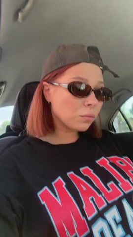 A young woman with short, reddish-pink hair, wearing a black t-shirt with "MALIBU BEACH" written in red and blue text, sunglasses, and a gray baseball cap backwards, is sitting in the passenger seat of a car. She is looking at the camera and smiling slightly. The interior of the car is visible behind her, including the car's headrest and part of the seat. The lighting suggests it is daytime, possibly with sunlight filtering into the car.