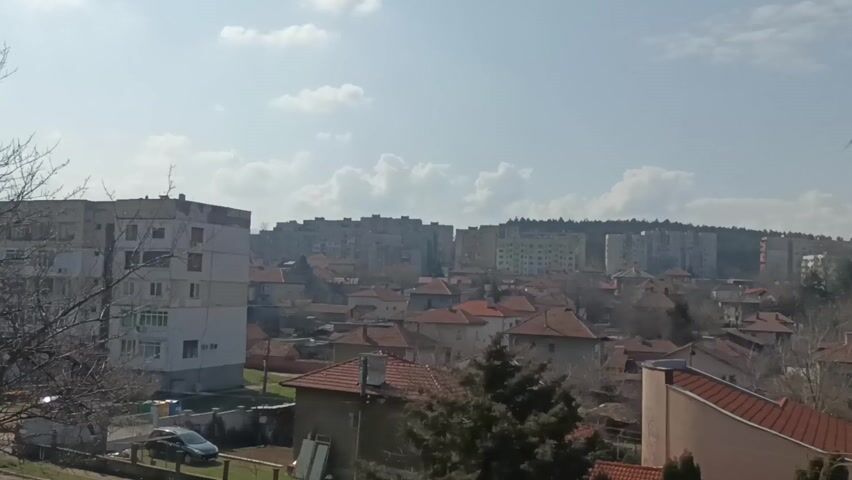 The video shows a cityscape on a sunny day with a few clouds in the sky. In the foreground are houses with red tile roofs and a few trees, some of which are bare. In the background, there are taller apartment buildings that appear to be on a hill. A car is parked in front of one of the buildings in the foreground. Audio of indistinct speech is also audible.