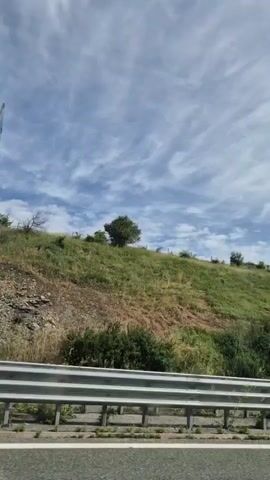 A video shows a hillside with green grass and some bushes. A flag, appearing to be white, green, and red, is flying from a flagpole on the top of the hill. The sky is blue with wispy white clouds. A metal guardrail is visible in the foreground, suggesting the footage was taken from a moving vehicle on a road. The camera is panning slightly to the right.