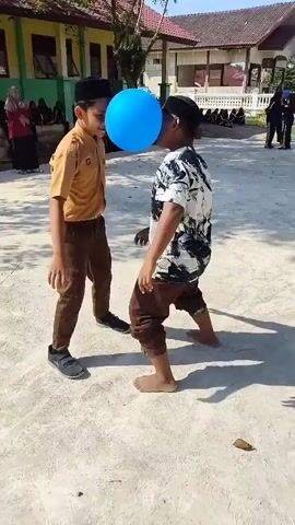 Two young boys are engaged in a game outdoors. The boy on the left, wearing a brown uniform with a light brown shirt and darker brown pants, and a black cap, stands with his feet apart, a blue balloon held between his forehead and the forehead of the boy on the right. The boy on the right, wearing a white t-shirt with a black abstract pattern and brown pants, also stands with his feet apart, barefoot. Both boys appear to be concentrating intensely on keeping the balloon in place. The background shows a building with a tiled roof and some trees, suggesting an outdoor school or community area. Other people are visible in the background, some seated and others standing. The ground is covered in light-colored sand or dirt. The sun is shining, casting shadows.