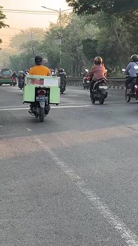 The video shows a street scene with a line of motorcyclists riding in the same direction. The motorcyclists are on a paved road with trees lining the sides. The sun is low in the sky, suggesting it is either early morning or late afternoon, and a warm, hazy light bathes the scene. Most of the motorcyclists are wearing helmets, and some are carrying items, such as a person in the foreground who has a light green box attached to the back of their motorcycle. The traffic appears to be moving at a moderate pace. The overall impression is one of daily commuting or travel on a busy road.