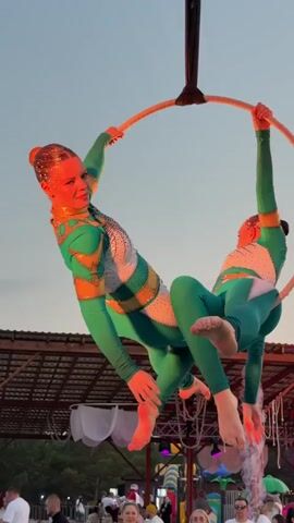 Two aerial acrobats perform on a hoop suspended from a rigging structure outdoors, possibly at an event or festival. The acrobats are dressed in matching teal and white costumes adorned with gold accents and glitter. One acrobat is positioned in front, looking towards the camera, while the other is behind and slightly above her. Smoke or mist is being emitted from below the hoop, creating a dramatic effect. In the background, there is a tent or canopy structure with lighting, a stage, and a crowd of people watching the performance. A drone is visible flying in the sky above the rigging. The sky appears to be transitioning from day to evening, with soft, warm lighting.