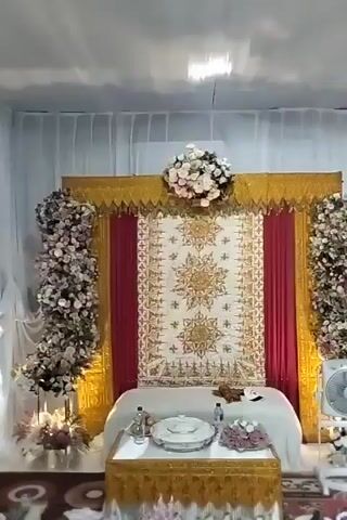 The video shows a ceremonial setup, likely for a wedding or similar event, with a decorated backdrop and a table laden with food. The backdrop features a large, ornate tapestry with intricate gold and white patterns, flanked by red curtains and gold trim. Floral arrangements, primarily in shades of pink and white, adorn the top of the backdrop and cascade down the sides. In front of the backdrop is a table covered with a white cloth, topped with several serving dishes and decorative items.

In the foreground, the floor is covered with a patterned carpet, and numerous plates of food are arranged in rows. The food appears to include various dishes, fruits like watermelon and oranges, rice cakes, and possibly boiled eggs. Some plates are also decorated with greenery. A white electric fan stands to the right of the setup. Towards the back of the scene, white curtains obscure the background, and a few people, mostly obscured, are visible in the distance, suggesting the event is in progress or about to begin. The lighting is bright, illuminating the decorated area.