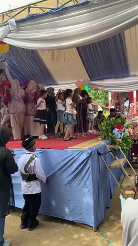 A group of children and adults are gathered on and around a stage, which is covered by a tent with blue and white fabric draped overhead. The stage has a red carpet. Balloons are attached to the tent poles. In the foreground, a young boy in traditional attire faces away from the camera, looking towards the stage. To the right of the stage, there are chairs and decorations, including flowers and greenery. The background appears to be an outdoor setting with trees and buildings visible. The event seems to be a performance or celebration.