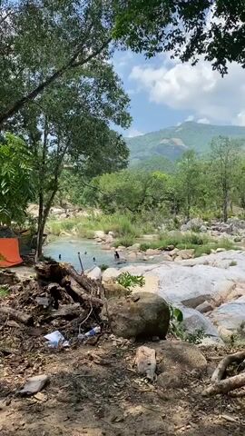 A group of people are seen swimming and playing in a shallow river. The river has a rocky bed and is surrounded by trees and lush vegetation. In the foreground, there is a pile of fallen logs and debris, with an orange tent partially visible. The background features rolling hills and a clear sky with scattered clouds. The overall scene depicts a natural outdoor environment with recreational activity.