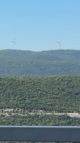 The video presents a landscape featuring two prominent green hills under a clear blue sky, with two wind turbines atop the more distant hill. The foreground contains a dense forest covering another hill. Beneath the foreground hill, a body of water is partially visible, bordered by pine trees. A gray metal railing appears along the bottom of the frame, suggesting the scene is being viewed from a bridge or elevated platform.