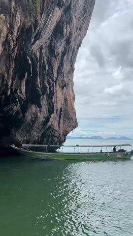 A long green boat with a canopy is floating in emerald green water, close to a massive, sheer cliff face. The cliff is dark brown with visible layers and texture. The boat has several people on it, some seated and one standing near the back. In the distance, more islands or landmasses are visible under a cloudy sky. The water is calm with small ripples.
