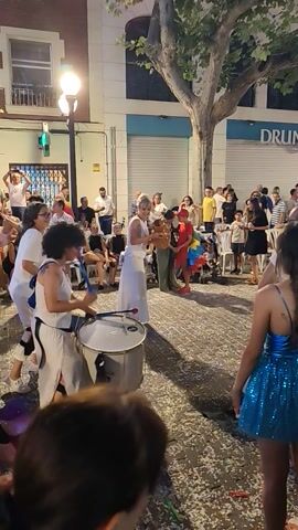 The video captures a lively street celebration with a crowd gathered. In the foreground, several people are visible, some with their backs to the camera. A person in the center is playing a drum, dressed in white with a white sash and black pants. To their right, another person in white also plays a drum. In the background, more people are observed, including a woman in a blue sequined dress. The street is decorated with confetti, and a tree with green leaves is on the left side. A building with windows and a sign that reads "FARMACIA Araceli" is also present. The lighting suggests it is either dusk or nighttime. The atmosphere appears festive and energetic.