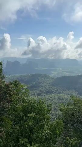 The video shows a scenic aerial view of a lush green valley with rolling hills and mountains. The sky is partly cloudy with bright sunlight illuminating the landscape. In the foreground, dense green foliage of trees frames the view. The middle ground features more forested hills and the distant background reveals a hazy panorama of more mountains and a vast, expansive landscape, possibly with some signs of human settlement or agriculture interspersed with nature. The camera appears to be panning slowly across the scene, capturing the vastness of the natural environment. No people or specific objects are identifiable within the frame. The overall impression is one of natural beauty and a sense of elevation, offering a wide vista.