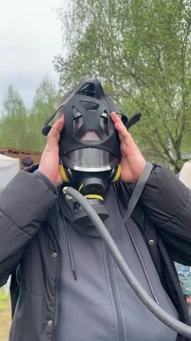 A man is shown from the waist up, putting on a black and yellow gas mask with a hose attached. He has his hands on the sides of the mask and is facing the camera. The man is wearing a dark jacket over a gray top. Behind him are other people and greenery, including trees and a white tent. The sky is overcast. A hissing noise is heard.