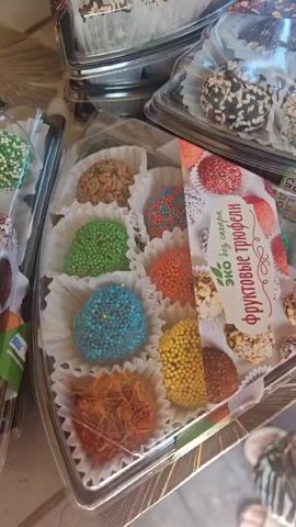 The video shows a close-up view of several transparent plastic containers filled with assorted chocolates or truffles. The truffles appear to be bite-sized and are arranged in individual paper cups within the containers. They come in various colors and textures, some are coated in sprinkles, nuts, or what appear to be dried fruits. One container has a price tag that reads "400." The background is slightly blurred but suggests a surface like a table. The lighting is adequate, allowing for clear visibility of the products. The overall scene is one of a display or selection of sweets, possibly in a store or market.