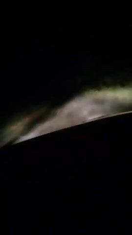 The video shows a first-person perspective of driving at night on a road. The headlights illuminate the road ahead, which appears to be wet or misty. Trees and vegetation are visible on the sides of the road, blurred due to the car's movement. The sky is dark.