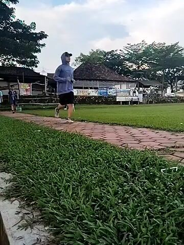 The video depicts an outdoor scene in Matesih, Indonesia, likely during the daytime under an overcast sky. A person wearing a light blue hooded shirt and dark shorts is shown jogging on a reddish-brown brick path. This path is bordered by a strip of lush green grass to the right and a narrow concrete drainage channel to the left.

In the background, several other individuals are visible further down the path. As the video progresses, three people are seen walking casually towards the camera. One person wears a light blue short-sleeved shirt, another a blue and white patterned shirt, and a third a yellow shirt.

The setting includes mature trees providing shade and several traditional-style buildings with dark, sloping roofs in the distance, suggesting a local community area, park, or leisure spot. Some of these buildings have signs or banners attached, but the text is not clearly legible. The overall ambiance is calm, with individuals engaged in light recreational activities.