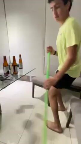 A young man is mopping the floor in a sparsely furnished room. He is barefoot and wearing a light green t-shirt and black shorts. The floor is white tile. A glass-topped table with three dark-colored beer bottles and a few other items sits to his left. Behind the table is a white wall. To his right are two gray chairs. The camera follows his movement and he briefly looks into the camera.