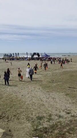 The video shows a beach where an event is taking place. Many people are walking on the sand towards the ocean. Some are wearing swimsuits or wetsuits, and many are wearing pink swim caps and carrying orange buoys. There is an archway marked "arena" and several blue tents set up near the water's edge. The sky is partly cloudy and the sea is visible in the background.