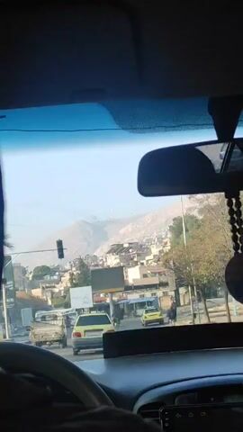 The video shows a view from inside a car driving on a road in what appears to be a city in Afghanistan. The sky is clear and hazy, with a mountain visible in the distance behind the buildings. There are several cars on the road, including a white truck, a yellow taxi, and other vehicles. A traffic light is visible on the left side of the road. A billboard displays Arabic script. Music is playing in the car.