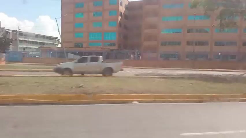 The video shows a street view with a large building in the background. The building appears to be several stories high with many windows that have a teal tint. A fence runs along the front of the building. There is a grassy area with a yellow curb in front of the fence. A white pole is visible in the grassy area. In the foreground, a road is visible with yellow lines marking the lanes. A motorcycle with two riders is seen passing by on the road. The riders are wearing helmets. The scene appears to be outdoors during the daytime, with sunlight casting shadows on the road.