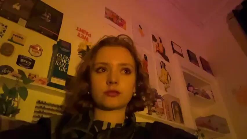 The video features a young woman with shoulder-length, curly brown hair indoors, lit by strong yellow-orange and pink-purple artificial light. She wears a dark top with a patterned collar and has eye makeup and red lipstick. Her expression is neutral as she occasionally looks towards the camera and then shifts her gaze around the room.

The setting appears to be a personal space, possibly a bedroom, with a wall decorated with various posters and shelves. Visible text on the wall includes an upside-down "WET PAINT" sign. On the shelves, a bottle labeled "GLENMORANGIE" is prominent, alongside round badges or coasters displaying "PETRO," "EFES," and an Irish shamrock symbol. Several artistic prints and illustrations, many depicting figures, adorn the wall. A green plant is visible on a lower shelf to the left. The lighting suggests it is evening or nighttime. There are no specific cues within the frame to indicate the location as Dundee, United Kingdom.
