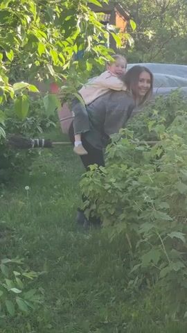 A woman with long brown hair, wearing a dark jacket and pants, is in a lush green garden with a small child on her back. The child, wearing a light-colored top and dark pants, clings to the woman. The woman is holding a dark-colored broomstick, positioning it as if she and the child are riding it. She bends her knees, rises, and moves slightly as if simulating flight on the broom. Both the woman and the child are smiling and looking towards the camera, appearing to laugh. In the latter part of the video, the woman holds a crumpled white object in her left hand. The garden features various green bushes, trees, and grass, with a wooden fence and a building partially visible in the background under sunlight.