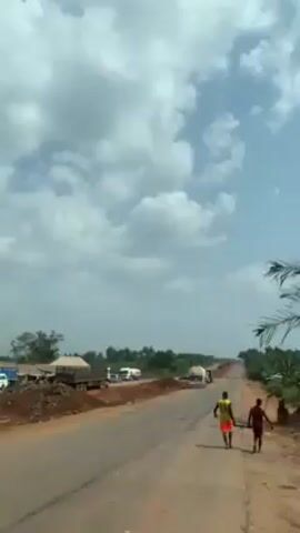 The video depicts a view from inside a moving vehicle, likely a car or truck, traveling down a paved road in a rural area. The sky is cloudy with patches of blue. In the initial shots, two people are walking along the side of the road. Several trucks and vehicles are parked along the roadside. The surrounding environment is characterized by trees and low-lying structures. The camera angle shifts to focus on the windshield wipers of the vehicle. The wipers are inactive.