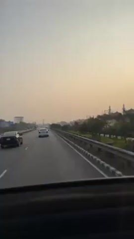 The video shows a view from inside a moving vehicle on a highway during daytime. The sky is a hazy, pale yellow, suggesting either early morning or late afternoon. Several vehicles are visible on the road, including a dark sedan in the left lane and a white sedan in the center lane. On the right side of the highway, there's a guardrail, followed by green vegetation and what appear to be buildings in the distance, some of which have tall, minaret-like structures. The overall scene gives the impression of an urban or suburban area on a major highway. A very loud wind noise is present during the recording.