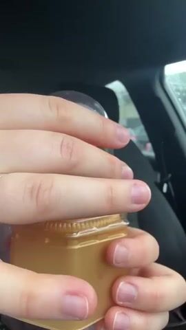 The video captures a person's hand holding a mango-shaped dessert inside a car. The dessert, housed in a golden, hexagonal container, displays an ombre color palette, transitioning from a vibrant reddish-pink at the top to a bright yellow at the base. The background reveals the interior of the car, featuring a black car seat and a window through which a rainy outdoor scene is visible. Towards the end of the clip, a woman brings the dessert to her mouth and licks it, with her face partially visible in a close-up shot. The scene appears to be set during a rainy day in Waldshut, Germany. No text is visible in the video.