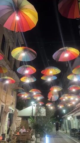 The video shows a vibrant night scene on a narrow street in Karditsa, Greece, decorated with numerous rainbow-colored umbrellas hanging overhead. Each umbrella has a light fixture beneath it, illuminating the street with a warm glow against the dark sky. On the left side of the street, an outdoor cafe or restaurant is visible, with several people seated at tables, enjoying the evening. Further down the street on the right, a person is seen walking away from the camera. Buildings with multiple stories line both sides of the street. Planters with trees or bushes are also present, adding to the urban ambiance. The overall atmosphere is lively and pleasant, typical of an evening out in a pedestrian area.