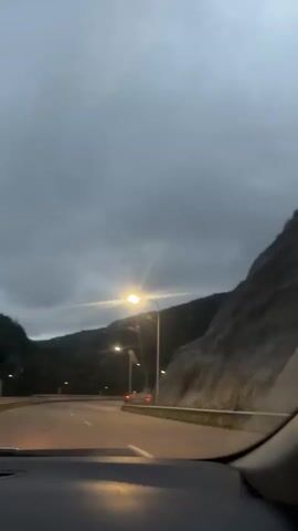 The video shows a view from inside a car driving on a highway at dusk. The road curves to the left, bordered by a rocky mountainside on the right. Streetlights illuminate the road, and the sky is overcast with dramatic clouds. Several cars are visible in the distance. The dashboard of the car is visible in the foreground. The lighting suggests that it is either early morning or late evening.