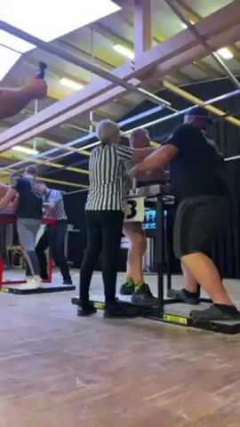 The video shows an arm wrestling competition. Two men are competing at a table, with a referee in black and white stripes standing between them. Other competitors stand at tables to the left. People are watching the match. The environment is a large room with a wooden floor and a high ceiling with metal beams. There is a sign with the number "3" on the table where the competition is happening.  The man on the right is wearing a black t-shirt and a baseball cap, while his opponent has a beard and is wearing a gray t-shirt and green socks.  A man in a black t-shirt and glasses is recording the event with his phone.